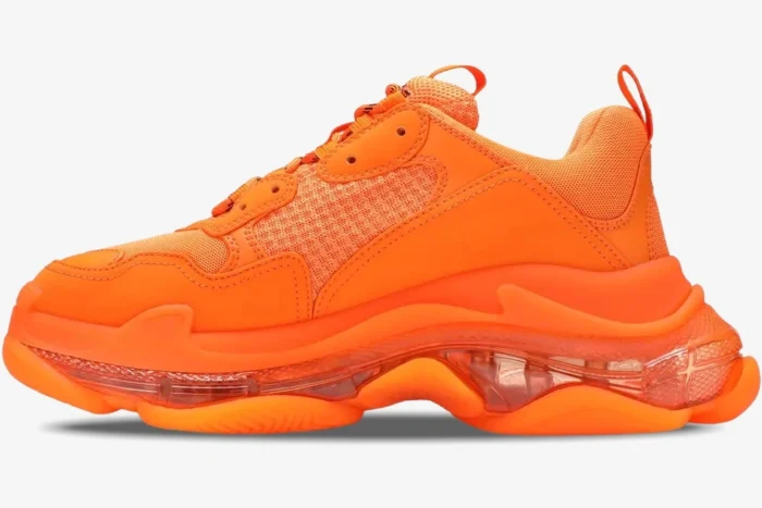 Side profile of a bright orange Balenciaga Triple S sneaker featuring a chunky clear sole.